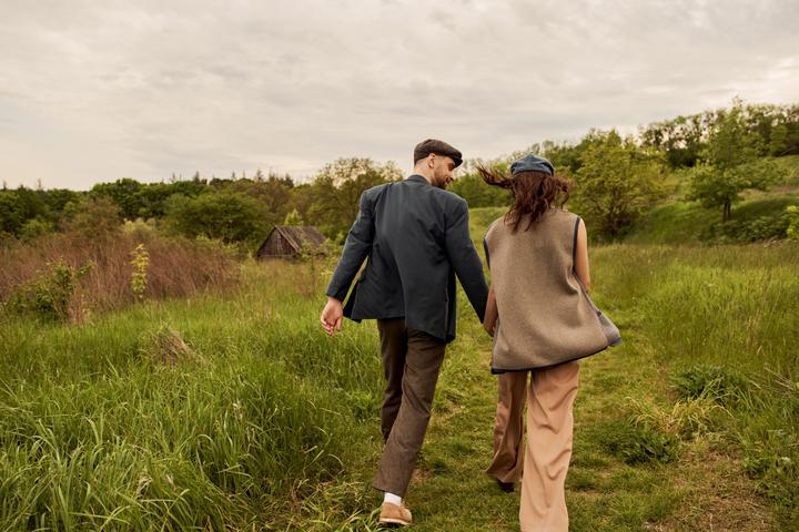 Bearded and stylish man in jacket and newsboy cap holding hand and looking at brunette girlfriend in